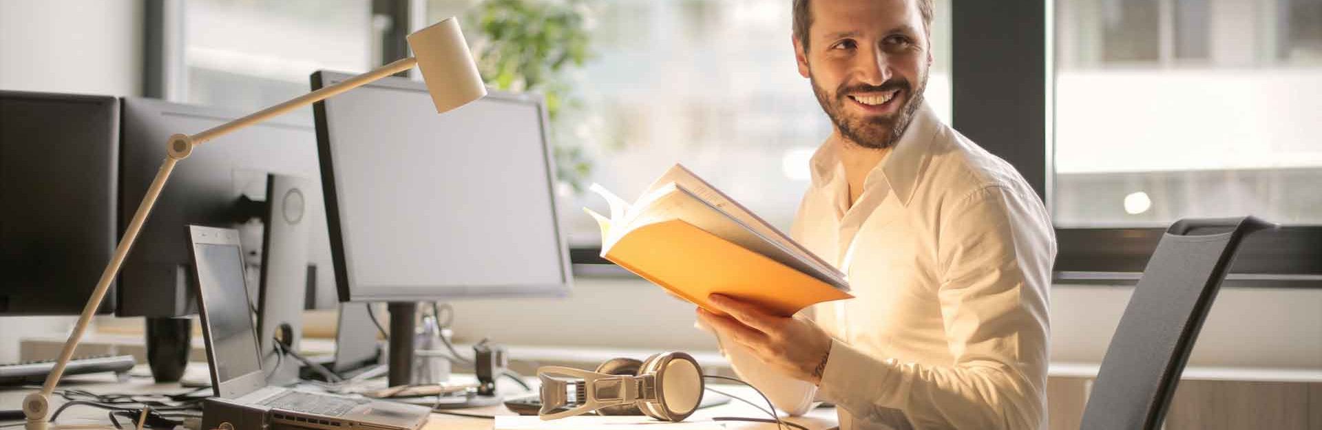 Smiling man sitting at computer desk while reading a book